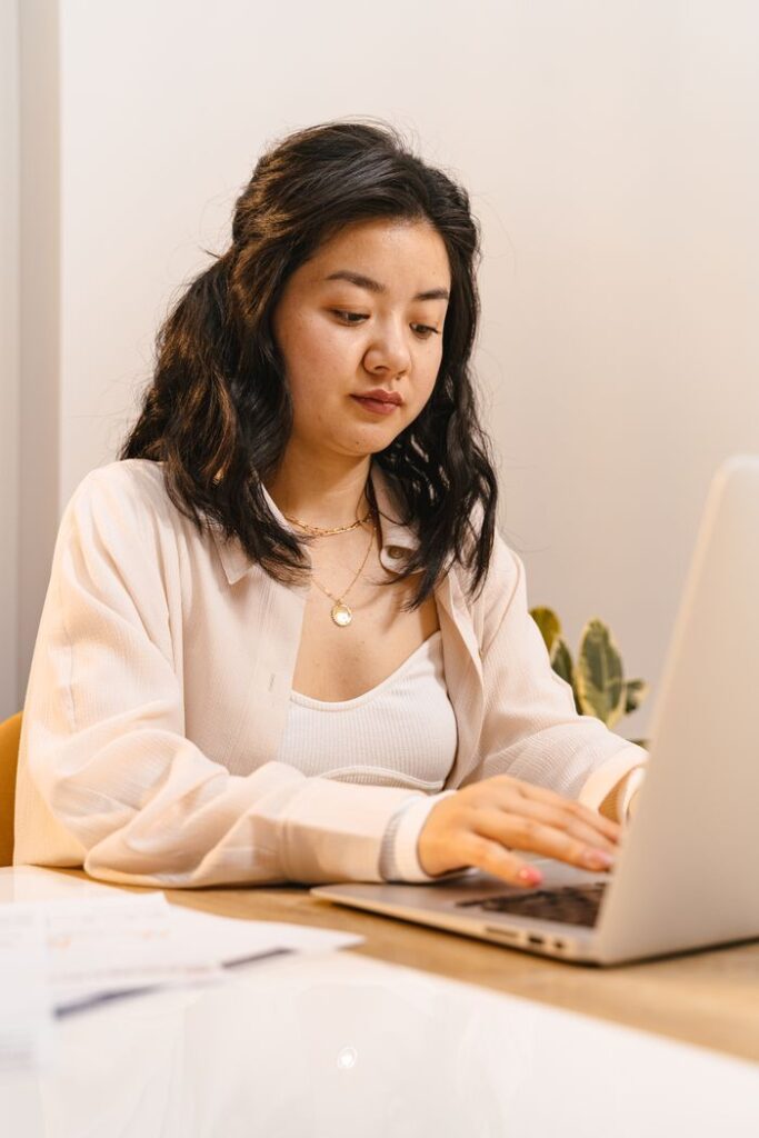 Asian woman concentrating on remote work using a laptop at a desk in a modern office space.